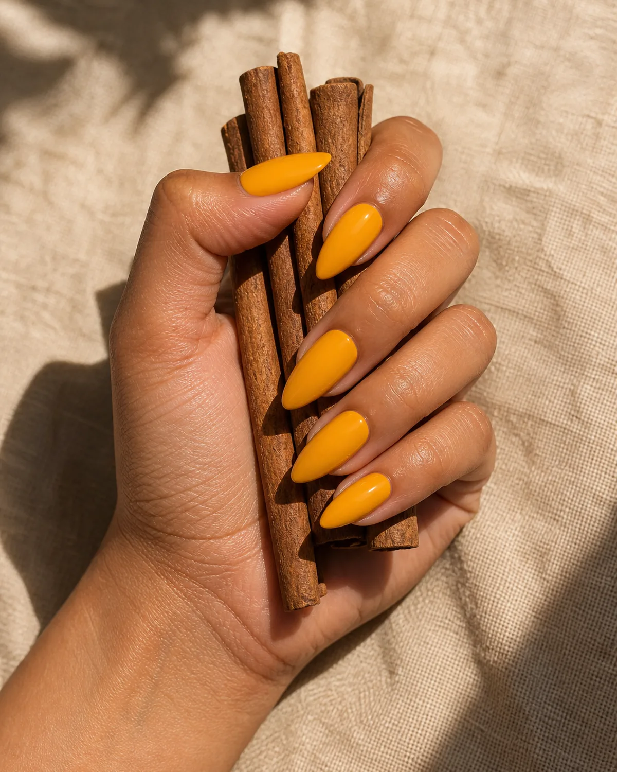Single hand with deep orange-tinged saffron yellow glossy polish on medium stiletto nails loosely holding cinnamon sticks on linen