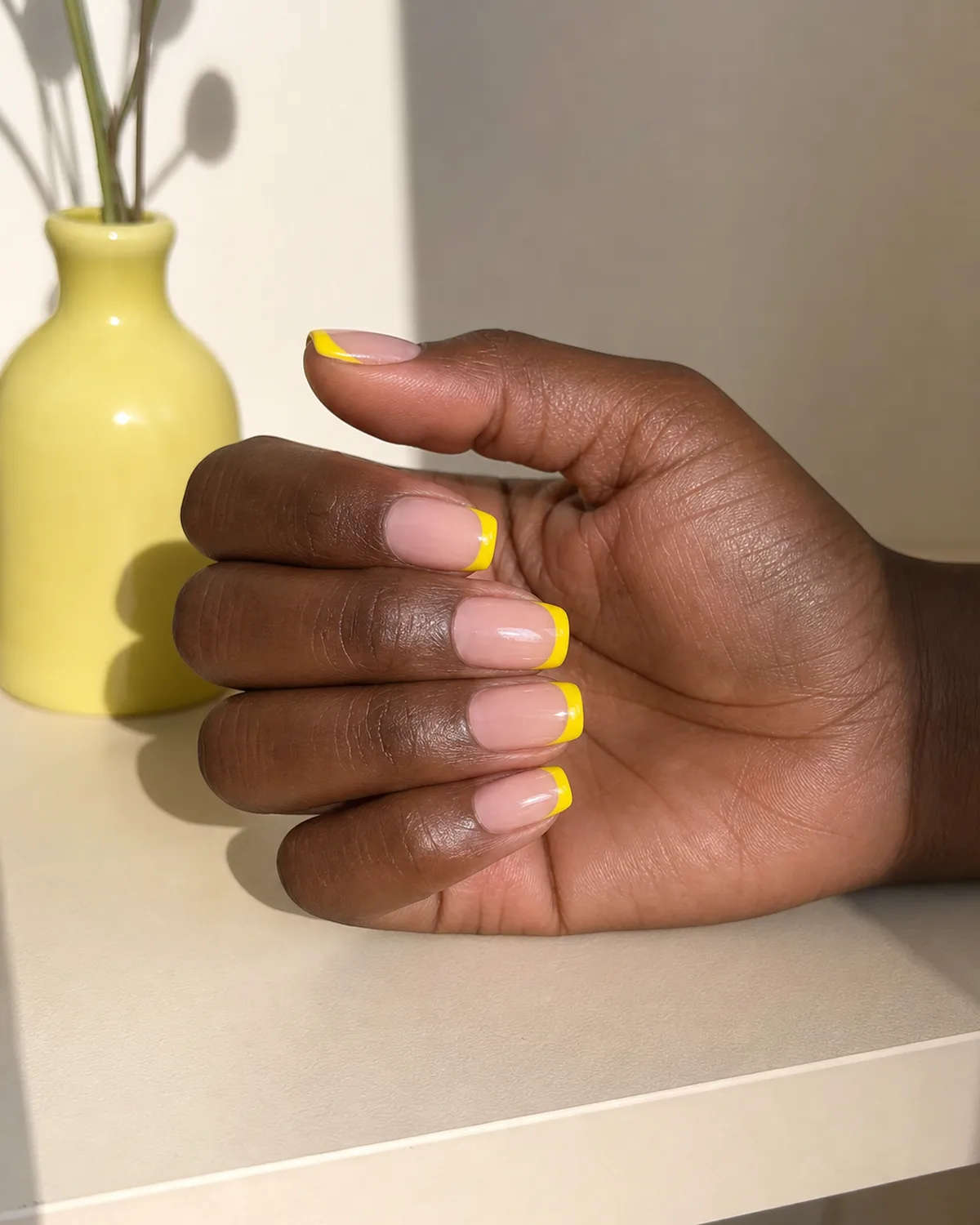 Single hand with bare natural base and opaque bright yellow French tips on squoval nails beside a small yellow ceramic bud vase on cream shelf