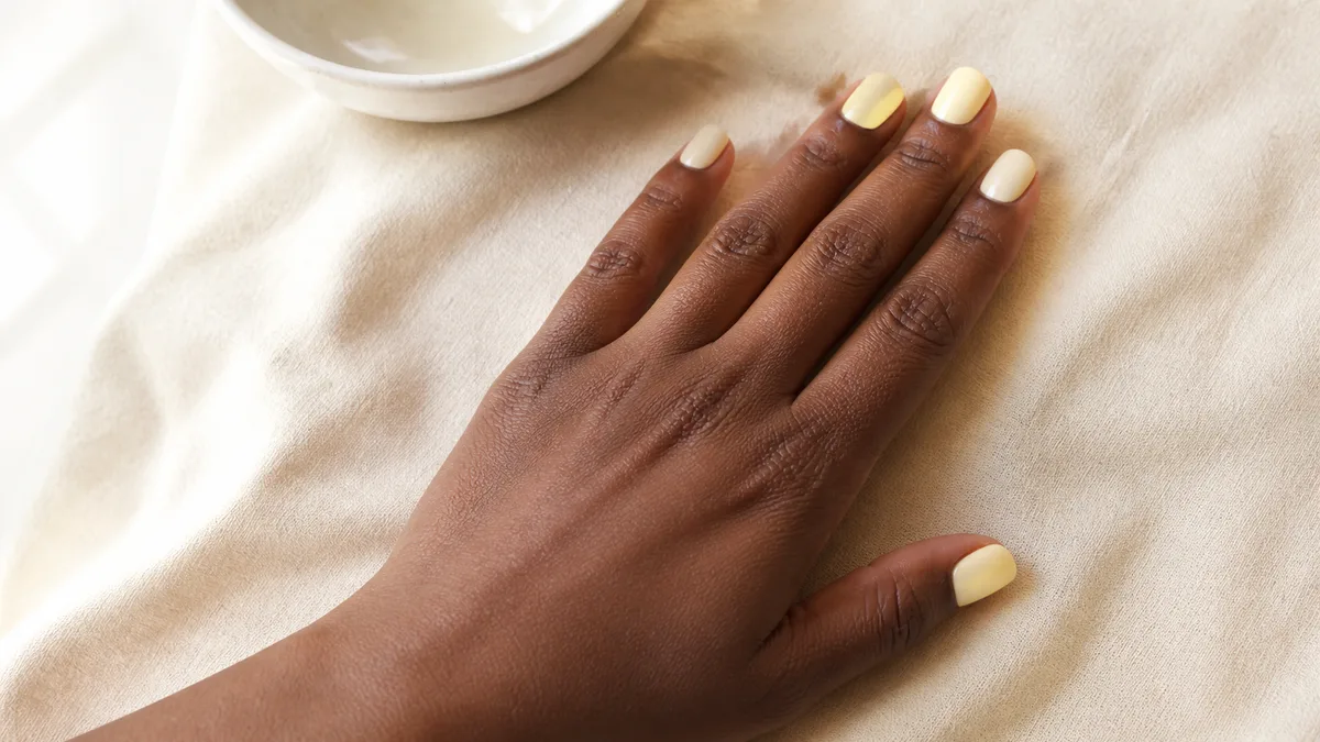 Single hand with soft butter yellow nails on short squoval shape lying flat on cream linen beside a small white ceramic bowl