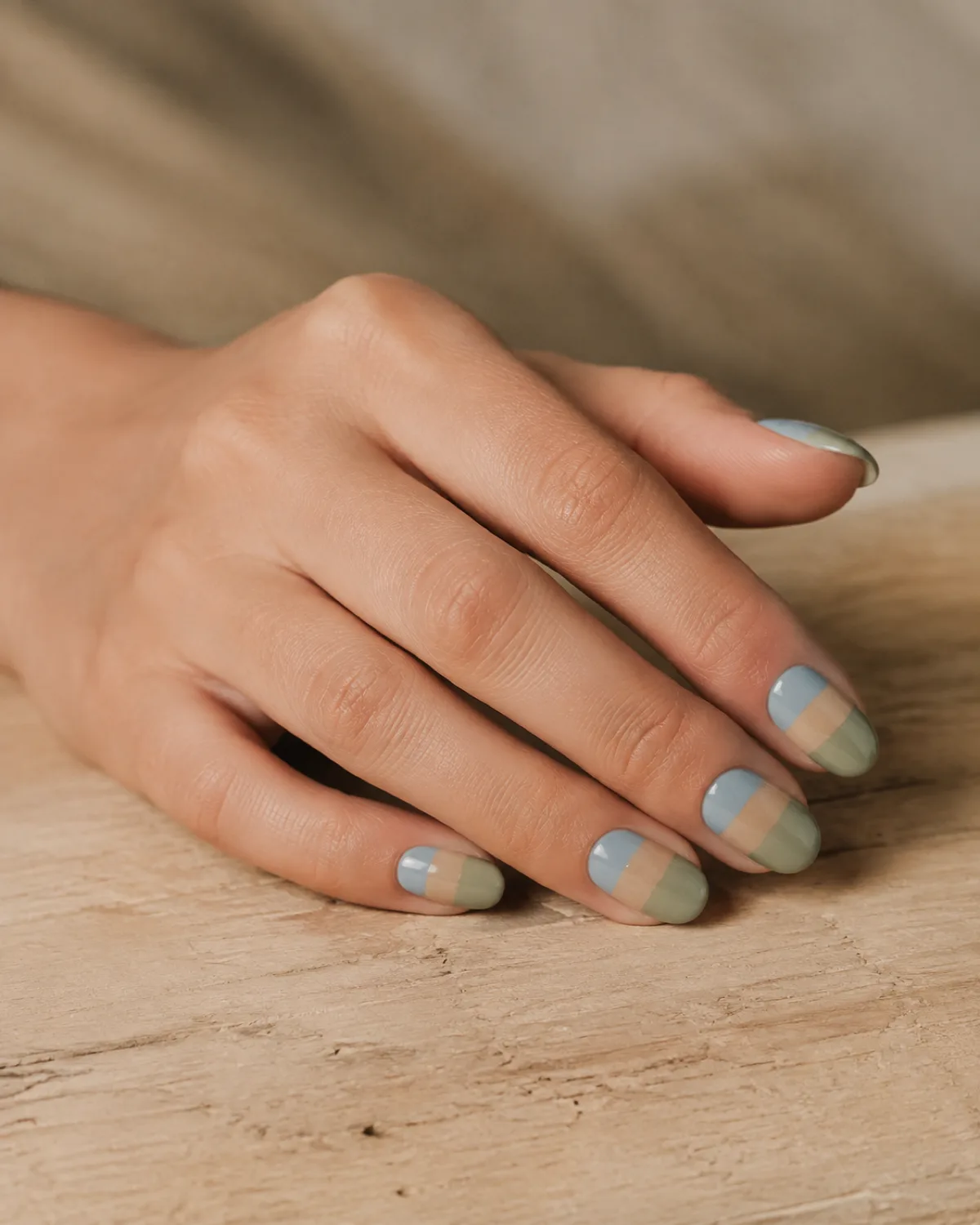 Single left hand with medium almond nails in muted abstract landscape color-field bands of sage sand and dusty blue on wood surface