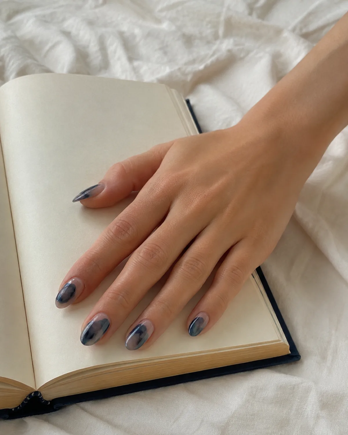 Single right hand with medium almond nails in ink-wash abstract of charcoal and indigo translucent patches on open book