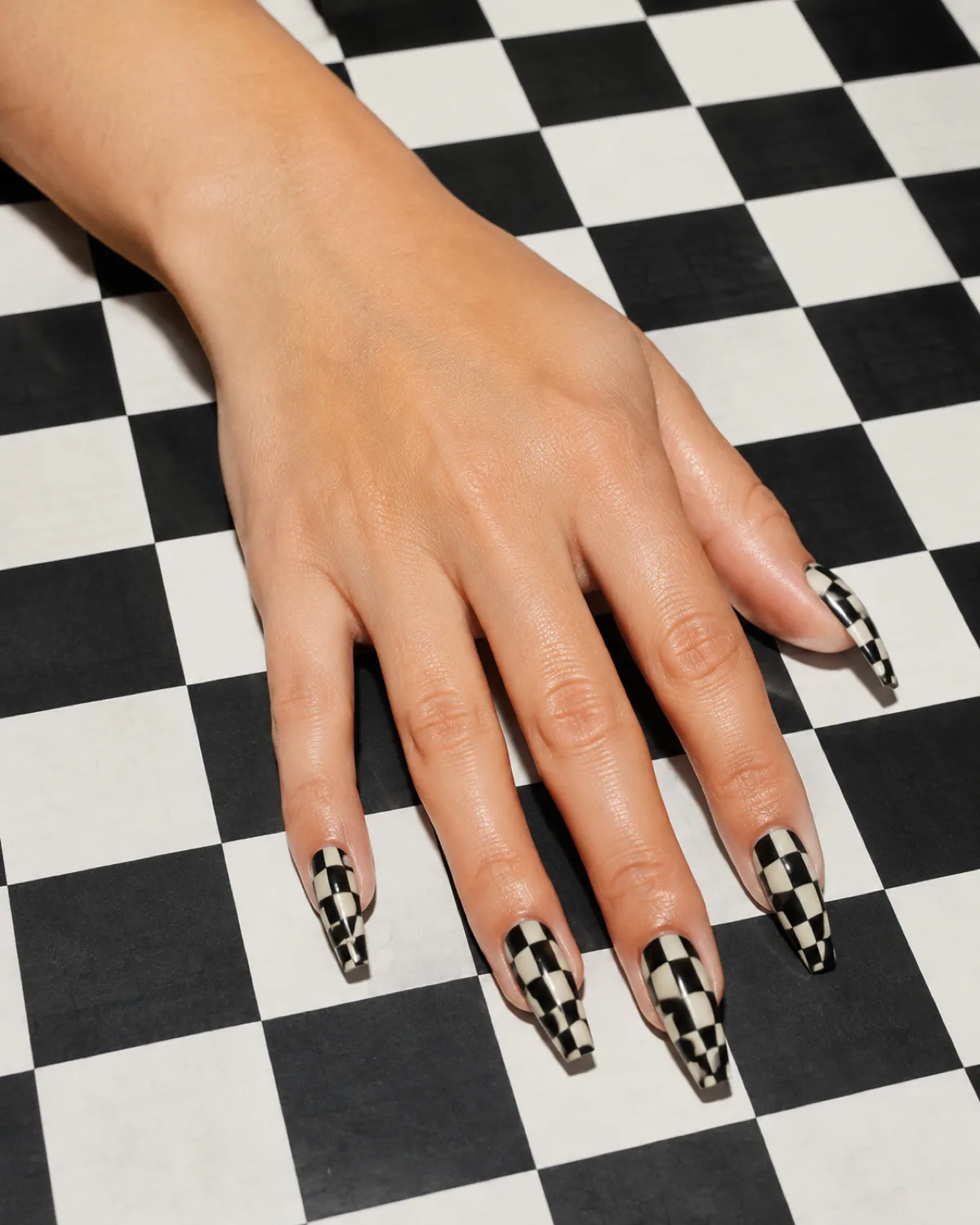 Single left hand with long coffin nails in warped checkerboard remix pattern in black and cream on checkered surface