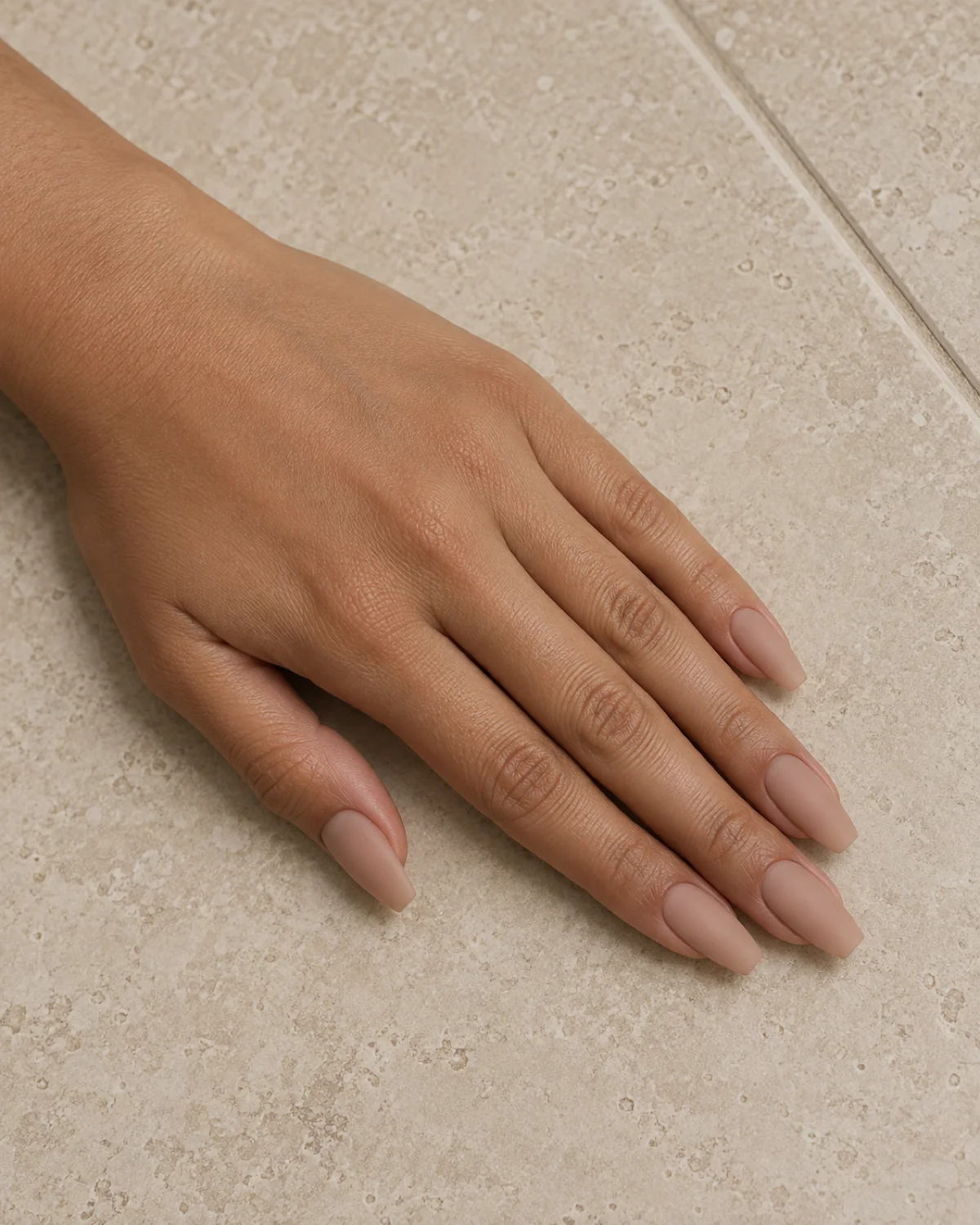 Single left hand with skin-tone nude matte polish on ballerina nails on a beige ceramic tile