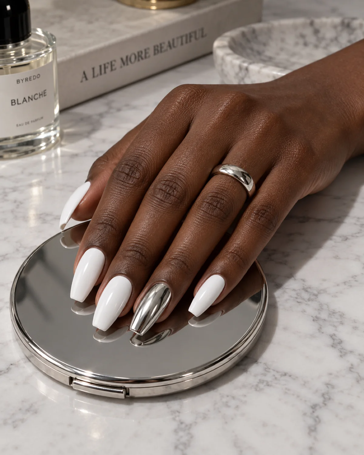 Single right hand with white polish and silver chrome ring finger on ballerina nails resting on a silver compact mirror