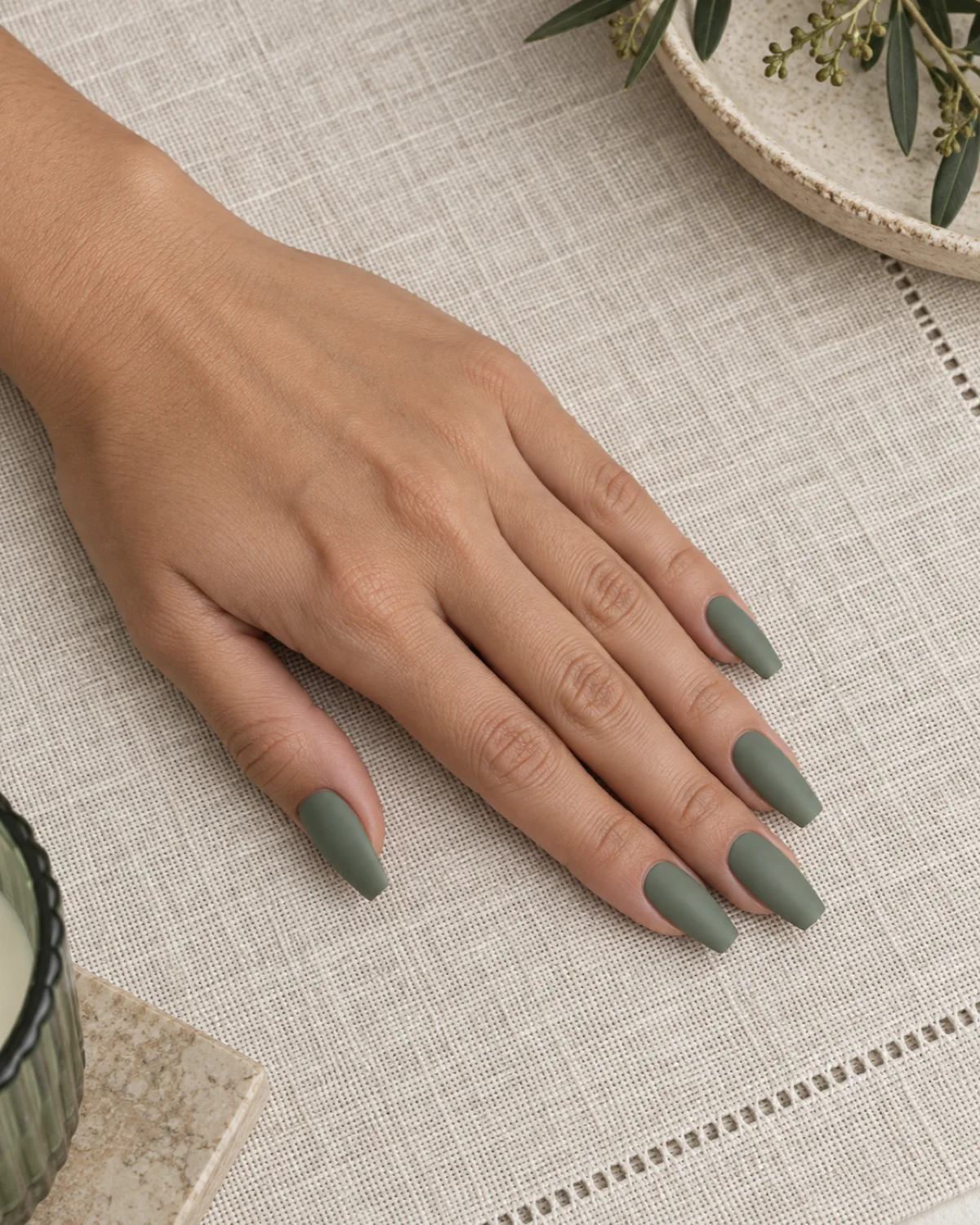 Single left hand with dusty sage-green matte polish on ballerina nails on a light linen placemat