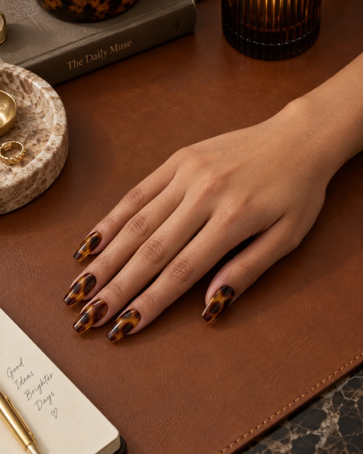 Single right hand with warm amber-and-brown tortoiseshell pattern on ballerina nails on a caramel leather desk pad