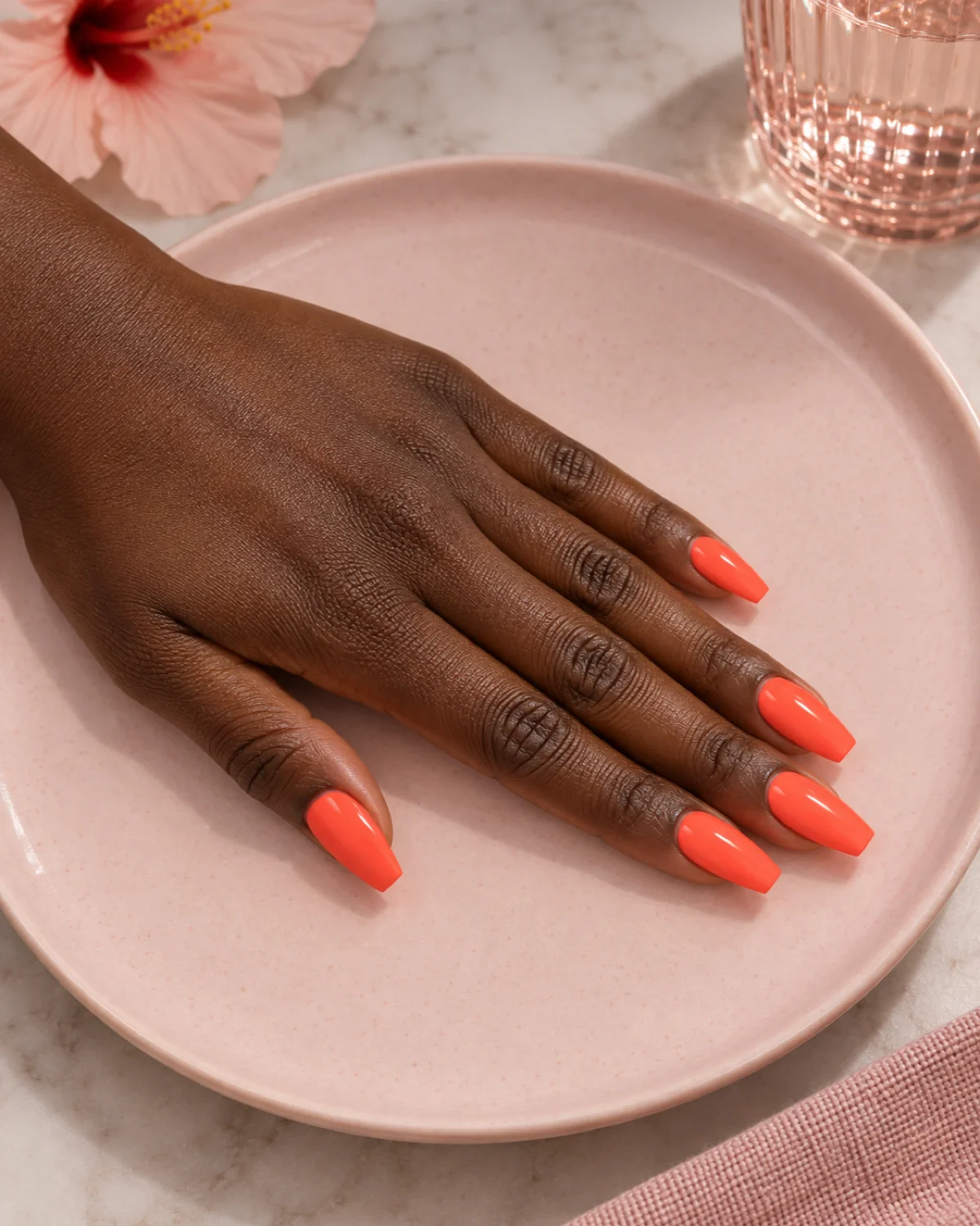 Single left hand with bright neon-coral polish on ballerina nails on a pale pink ceramic plate