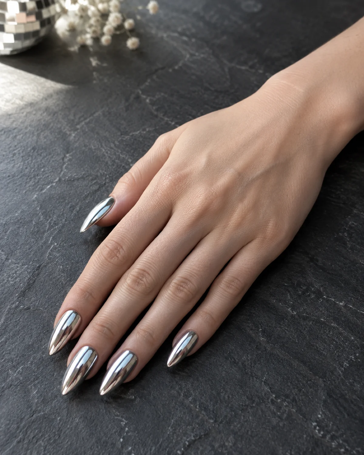 Single right hand with silver disco mirror birthday nails on stiletto shape resting on dark grey slate