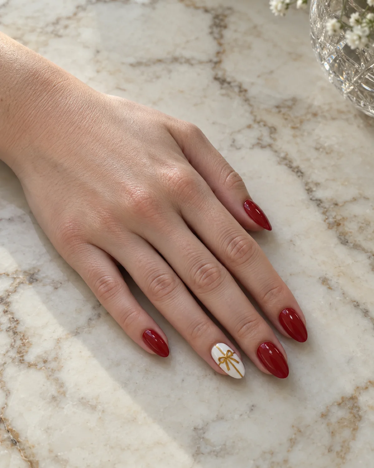 Single left hand with gift-wrap bow accent birthday nails on cherry red almond shape resting on cream marble