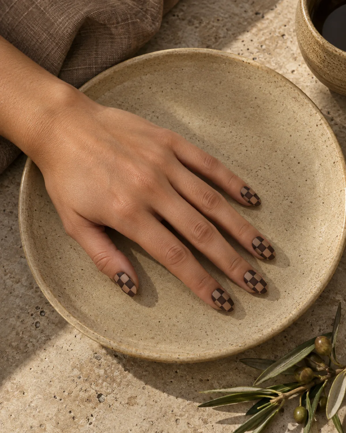 Single left hand with warm nude and brown matte checkered oval nails on a sand ceramic plate
