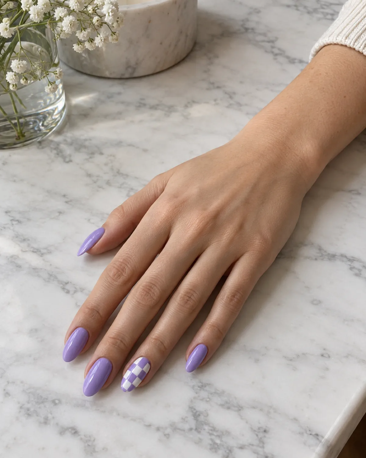 Single right hand with solid lilac almond nails and lilac-and-white checkerboard on ring finger on white marble