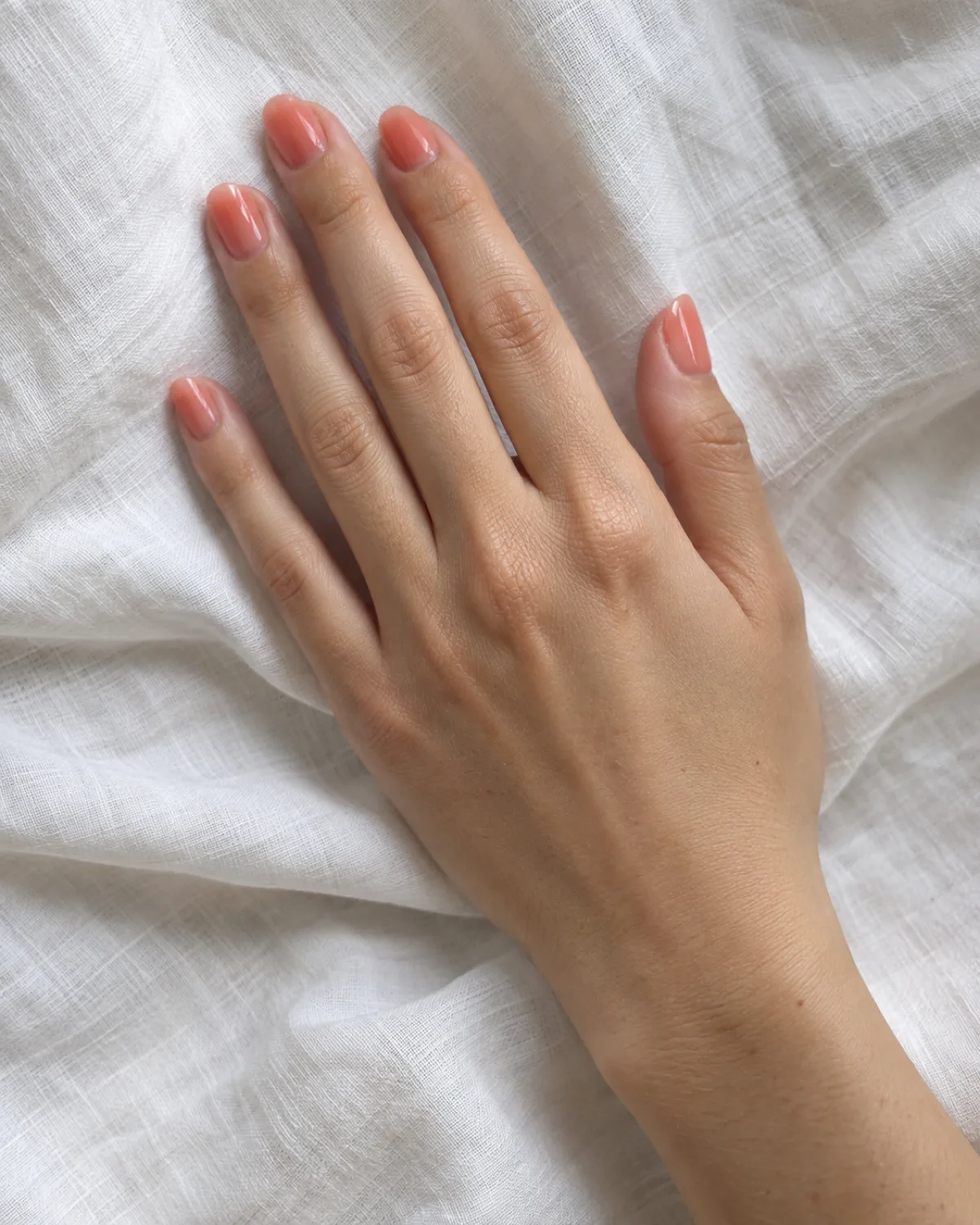 Single right hand with round nails in sheer coral jelly polish lying relaxed on its side on white linen cloth