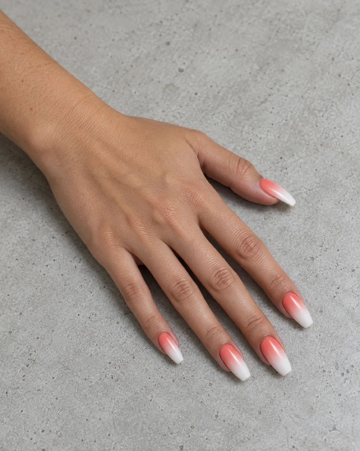 Single left hand with coffin nails in coral-to-white ombré gradient on light grey concrete surface