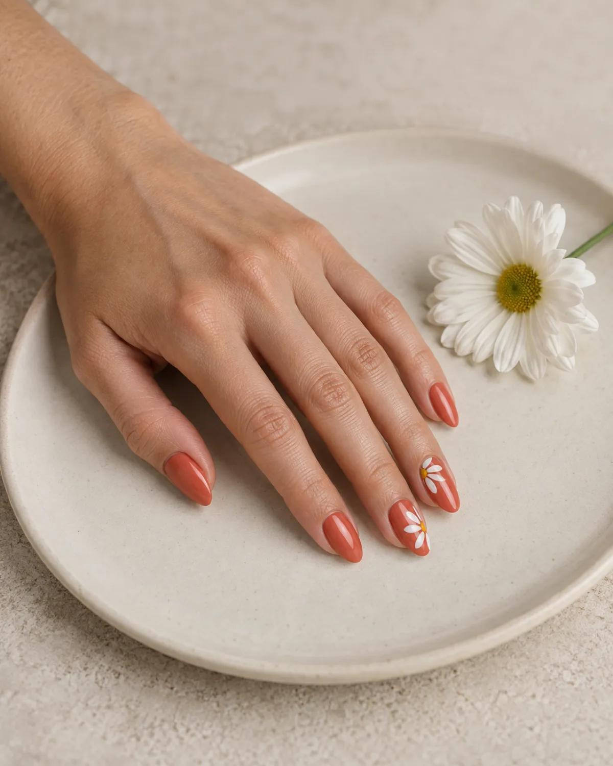 Single left hand with almond nails in warm coral base with white daisy petal art on accent nail resting on cream ceramic plate with a white daisy