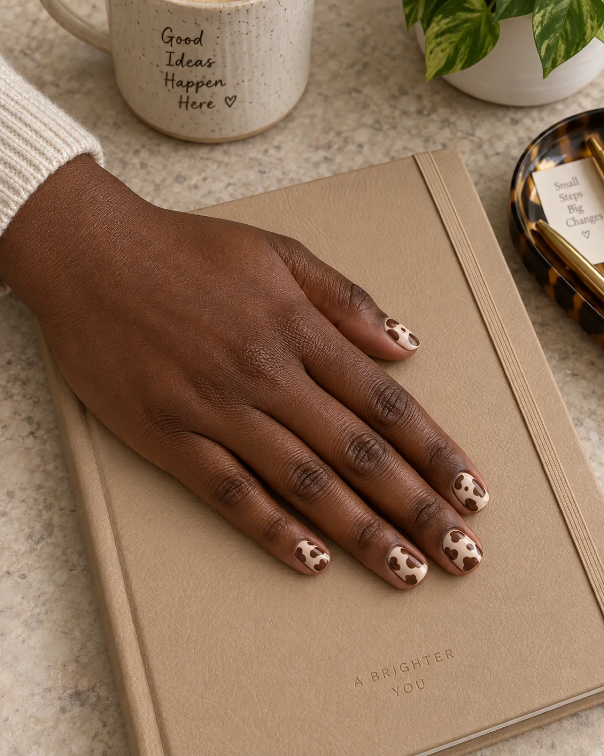 Single left hand with brown cow print nails on short square shape resting on a light-tan leather notebook