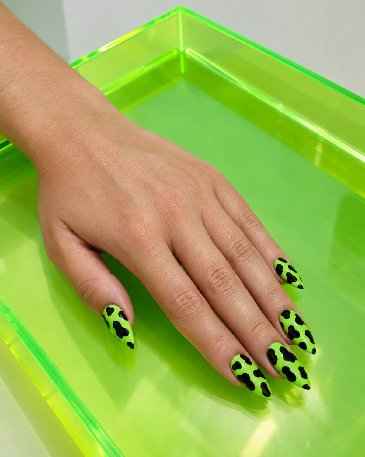 Single left hand with neon green and black cow print nails on stiletto shape resting on a neon-green acrylic tray