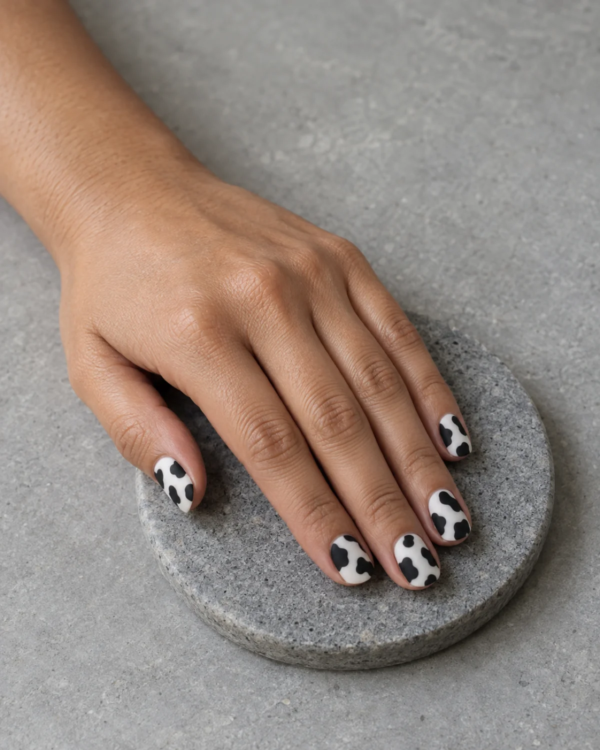 Single left hand with matte black-and-white cow print nails on short round shape resting on a flat gray stone coaster
