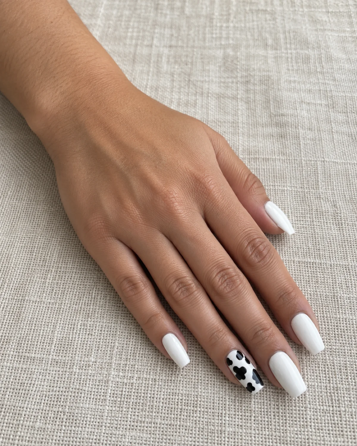 Single left hand with white coffin nails and cow print accent on ring finger resting on a cream-colored linen placemat