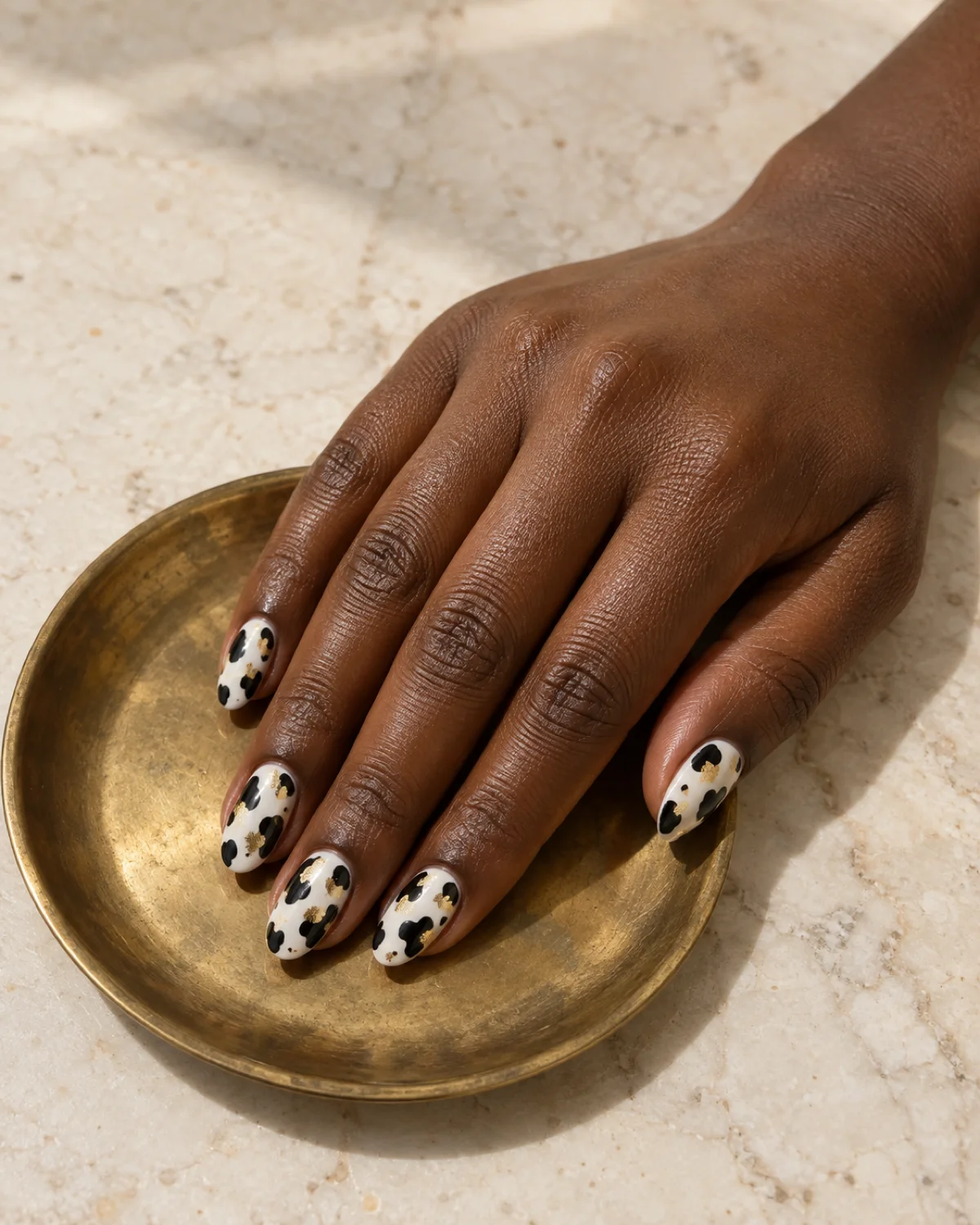 Single right hand with black-and-white cow print nails with gold foil flecks on almond shape resting on a small brass dish