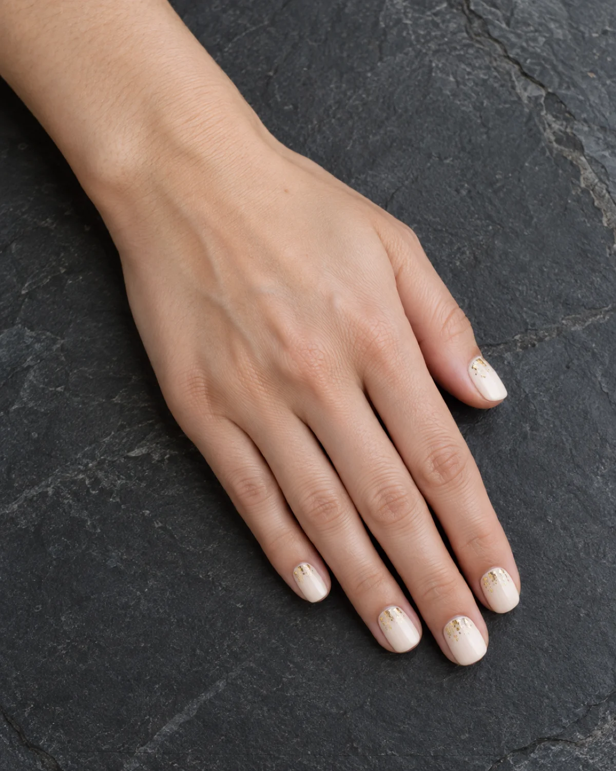 Single left hand with ivory and gold flake elegant nails on short squoval shape resting on dark slate tile