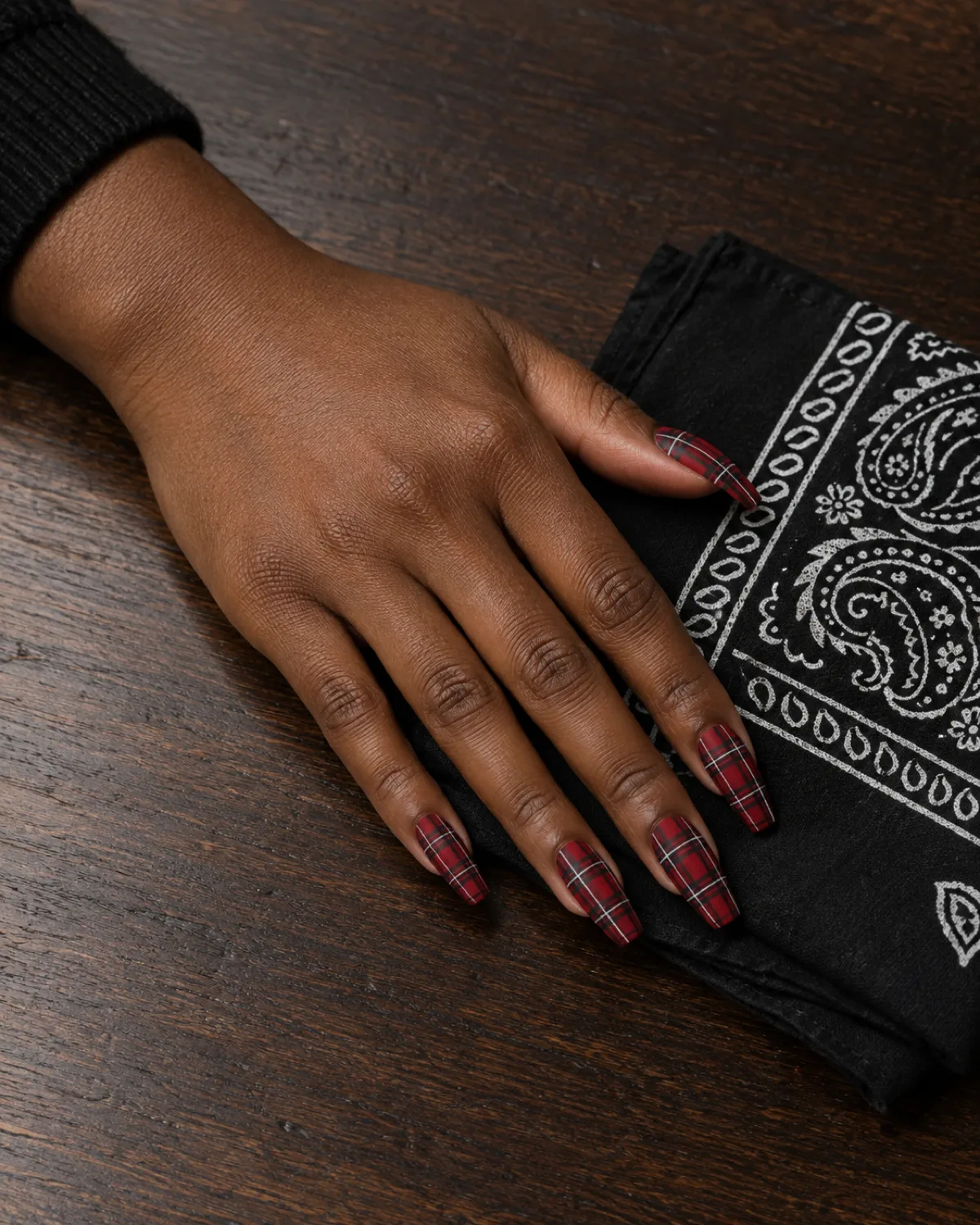 Red tartan plaid pattern coffin nails with black and white lines on deep skin tone hand beside folded bandana on walnut table
