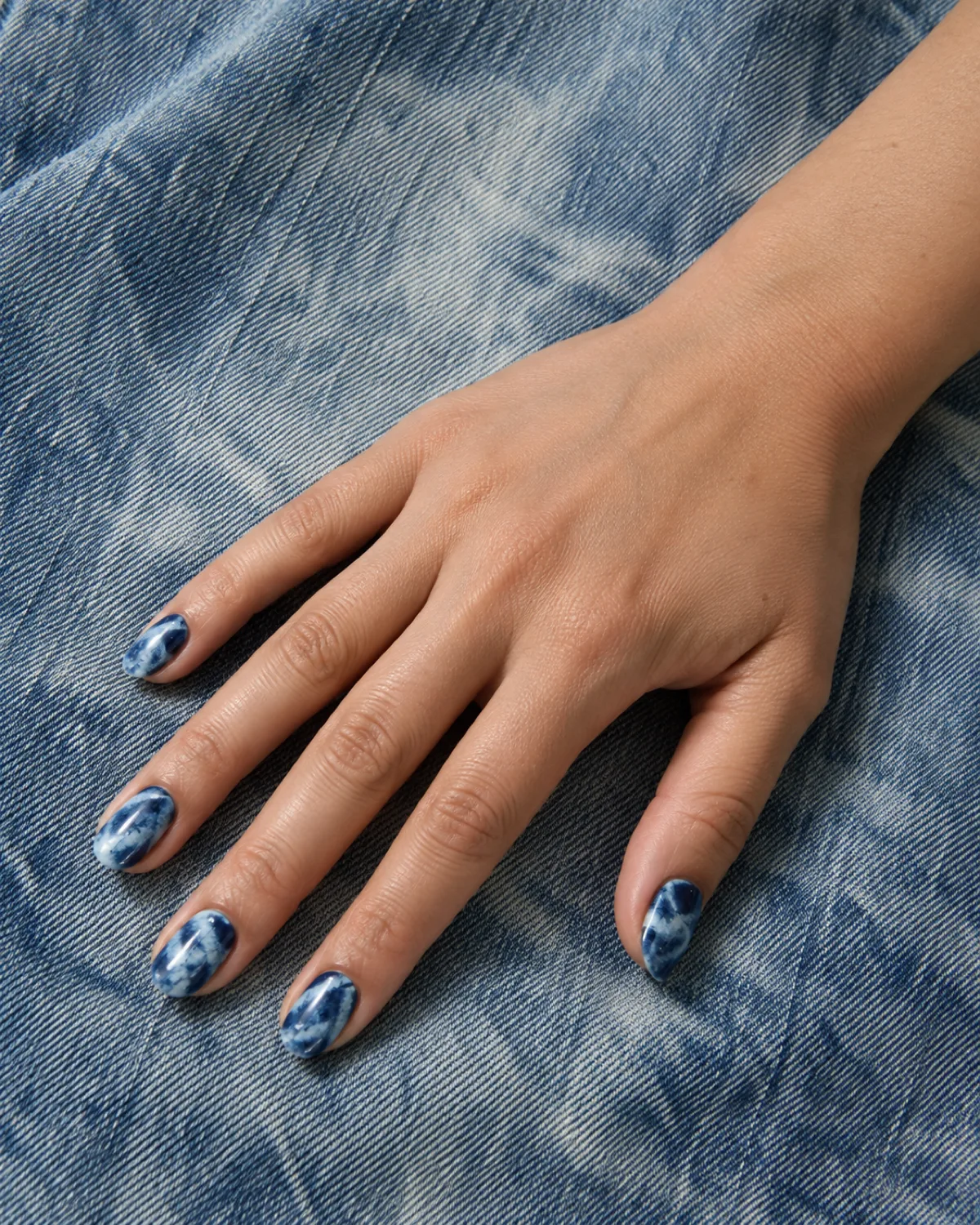 Acid-wash tie-dye nails in indigo and pale blue swirl on oval shape against faded denim fabric