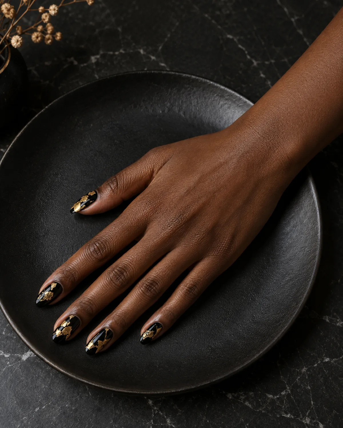 Glossy black almond nails with torn irregular gold foil pieces on deep skin tone hand on matte black plate