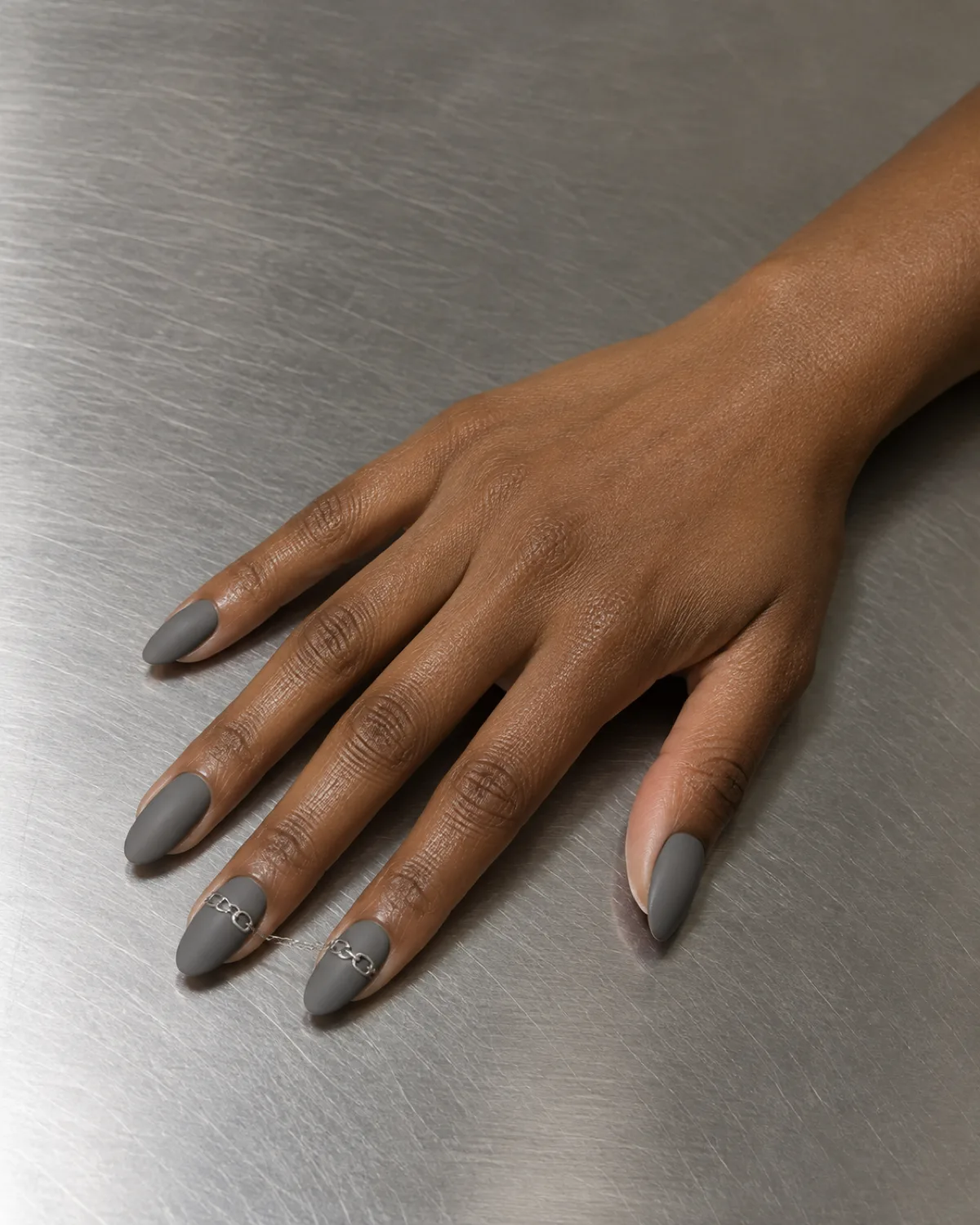 Matte grey medium almond nails with thin silver chain-link line across middle and ring fingers on brushed steel surface