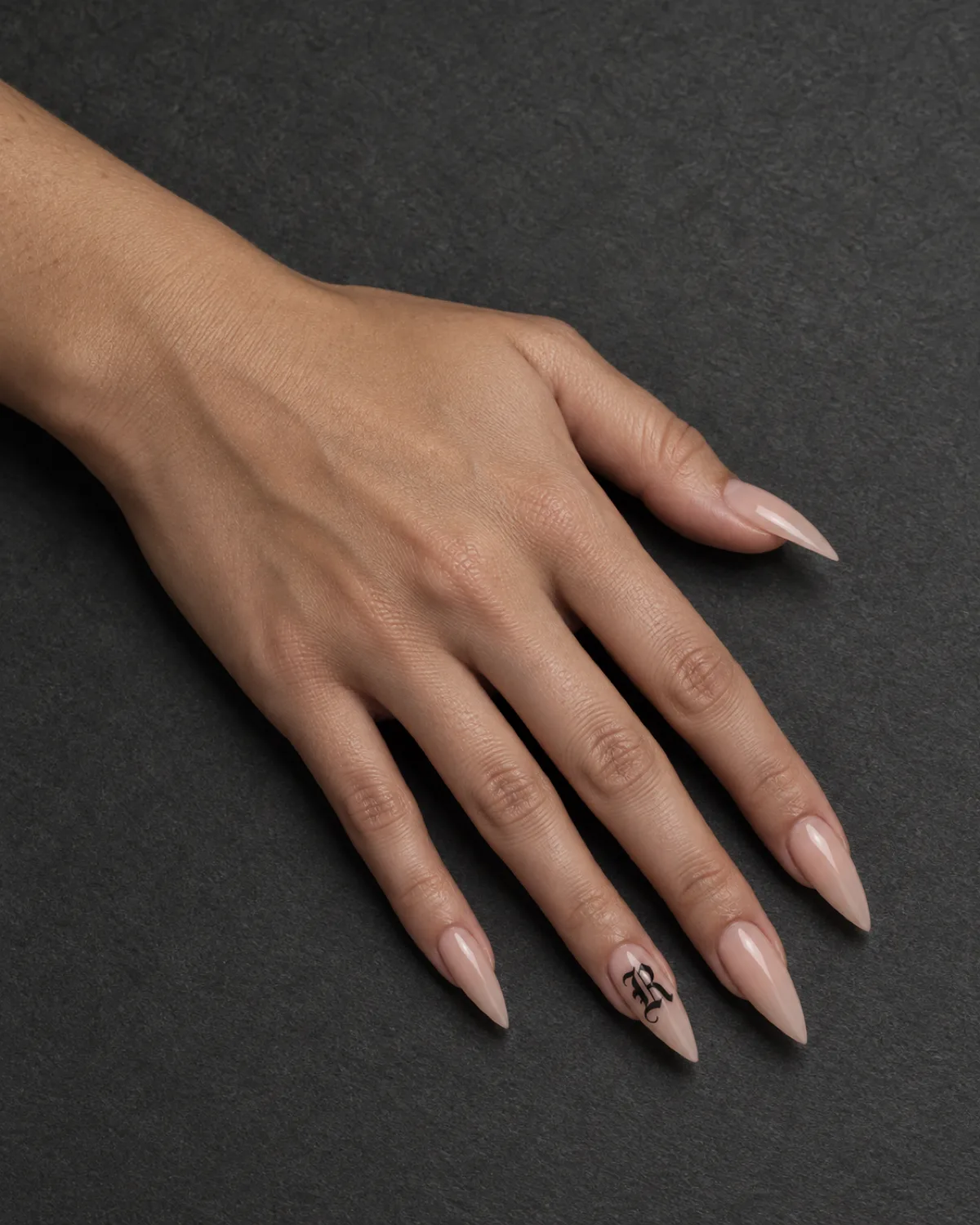Nude stiletto nails with ornate gothic blackletter initial in matte black on ring finger on dark grey textured paper