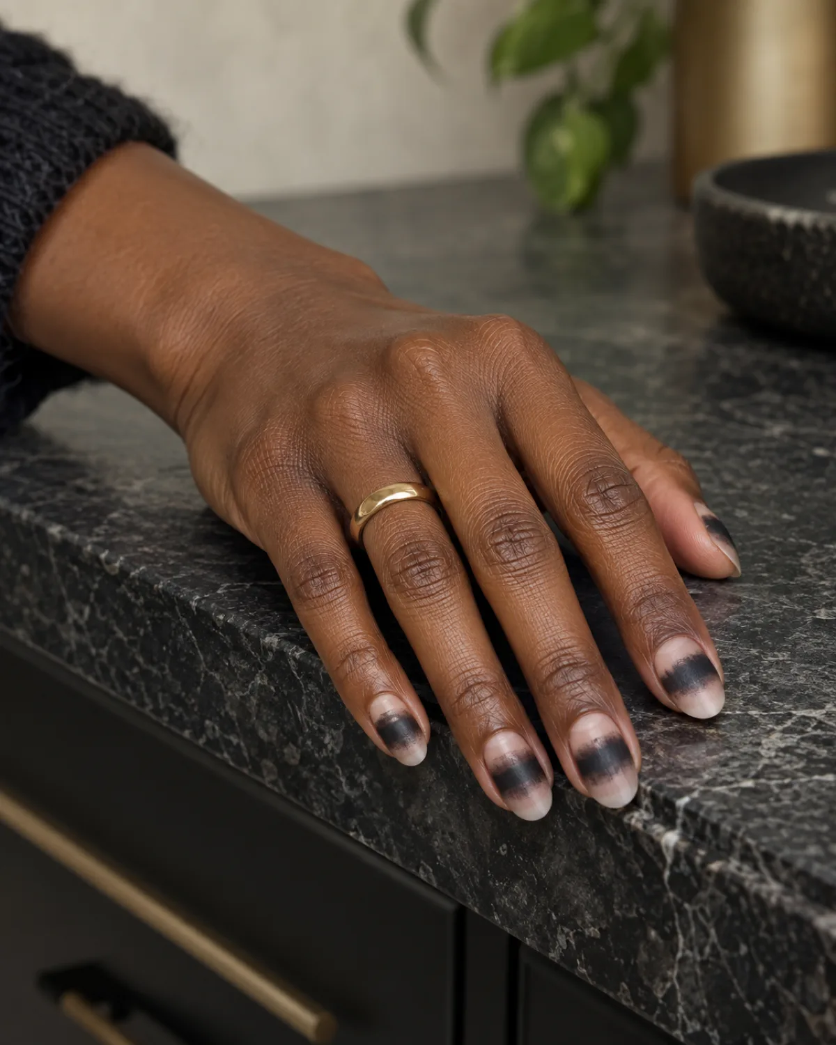 Black pigment smudged in a band across oval nails creating smoky eye effect on deep skin tone hand on stone countertop edge