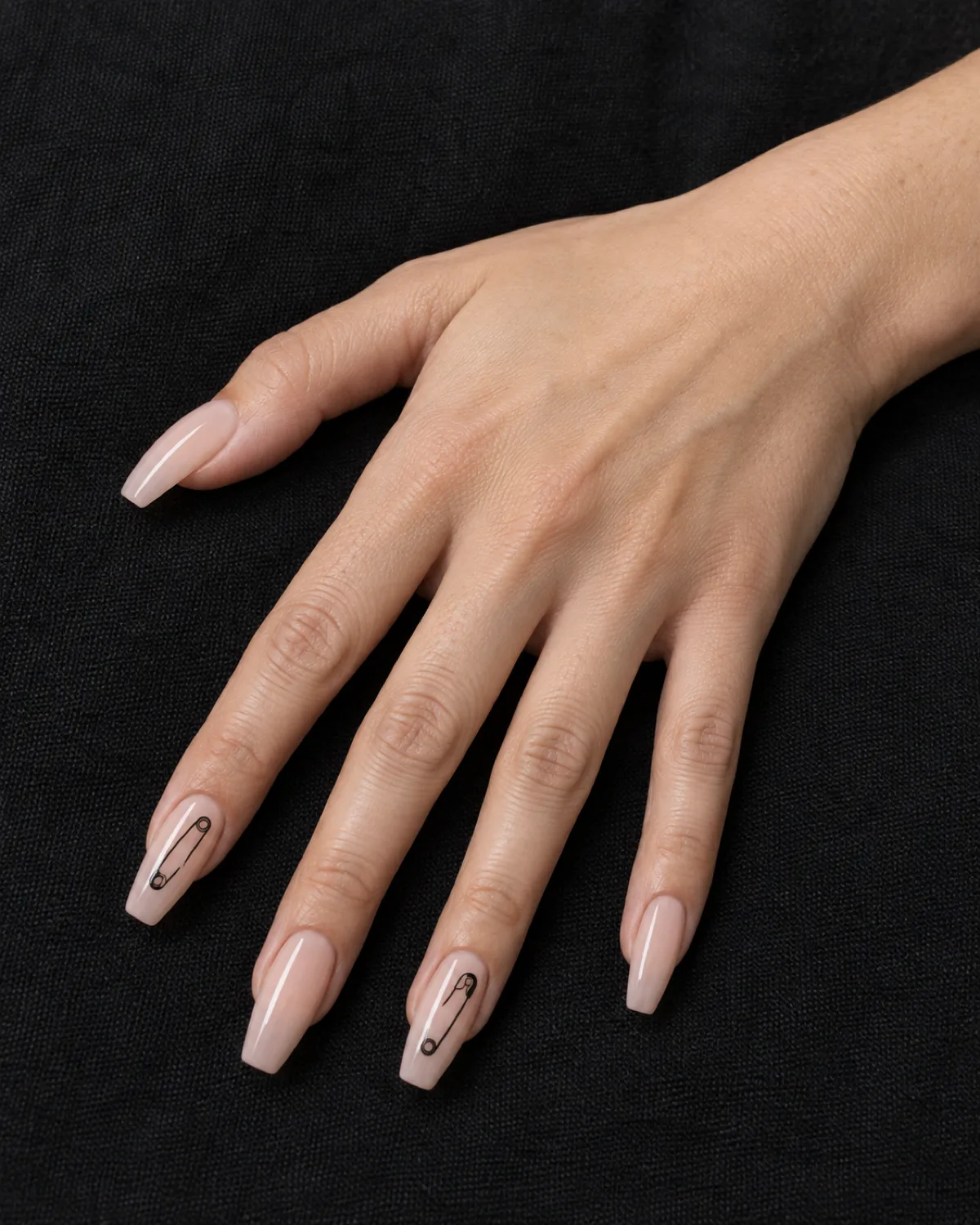 Thin black safety pin line art on nude coffin nails on black cotton canvas background