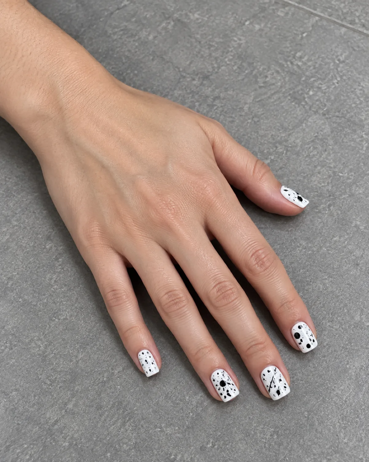 White square nails with random black paint splatter dots and flicks on grey ceramic tile