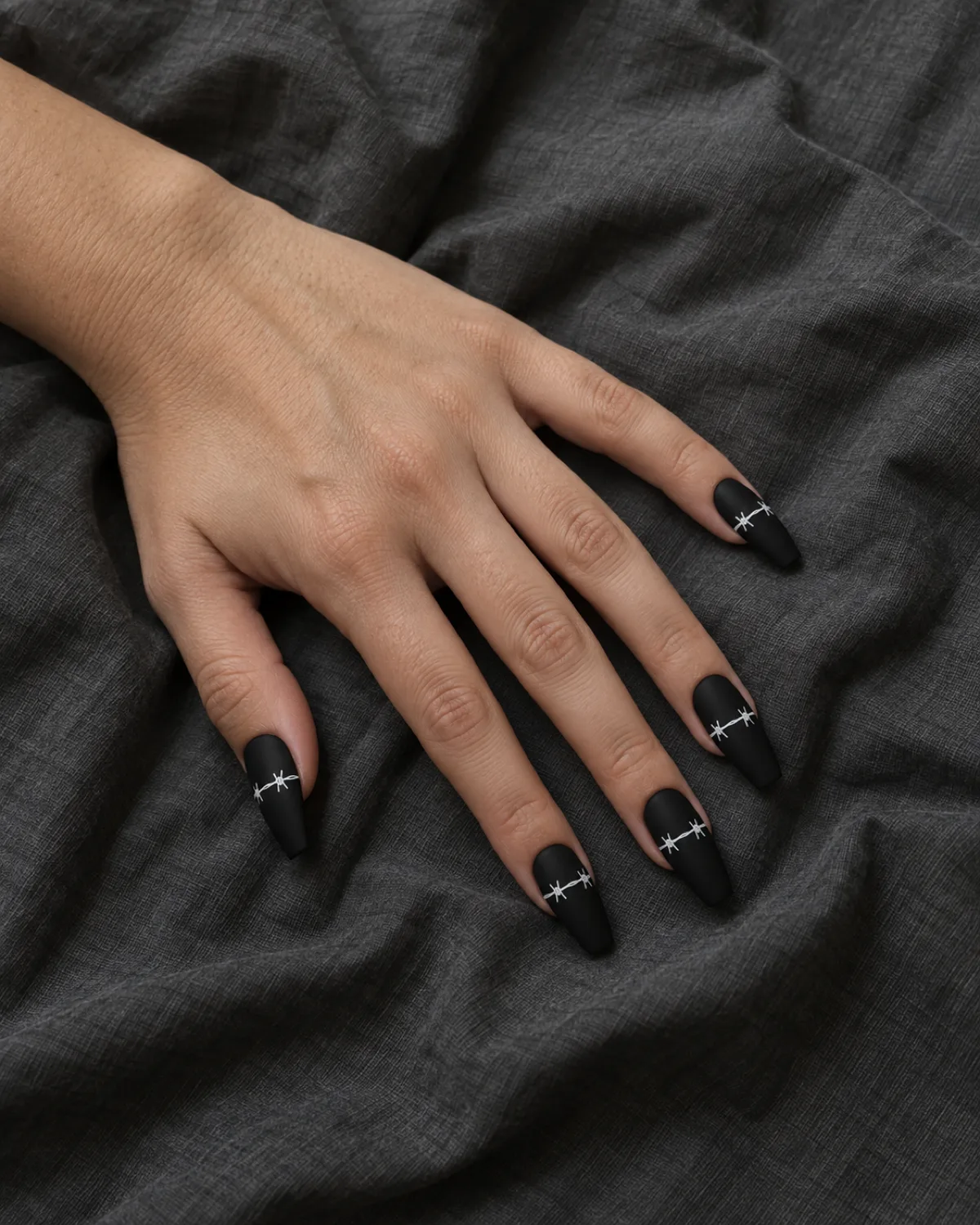 Matte black coffin nails with delicate white barbed-wire line drawn across all five nails on dark grey crumpled fabric