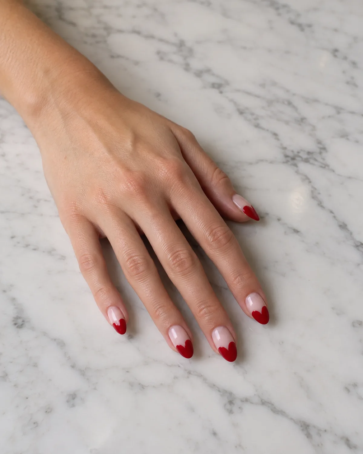 Single left hand with almond nails featuring red heart-shaped French tips on sheer pink base, on white marble countertop