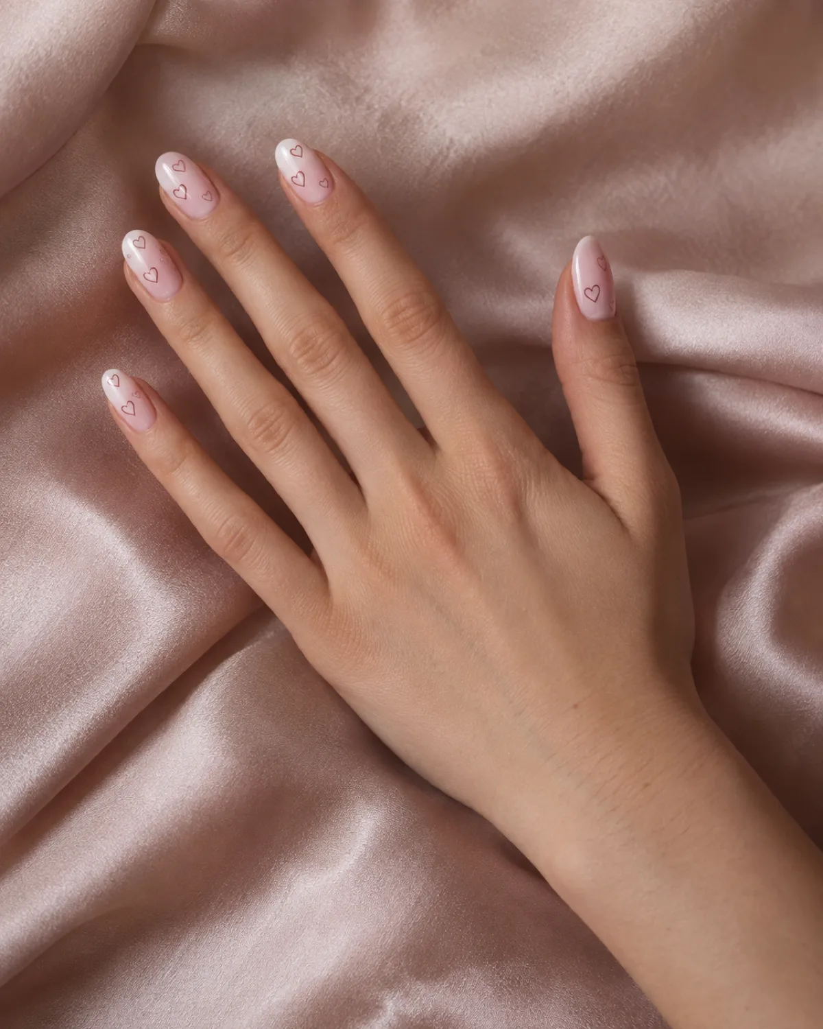 Single right hand with round nails featuring pink-to-white ombré base and tiny outlined hearts scattered at different angles on each nail, resting on blush satin