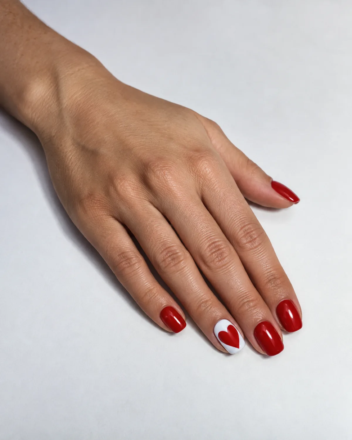 Single left hand with squoval nails, all solid cherry red except ring finger showing a large cherry red heart filling the entire white nail plate, flat on white paper