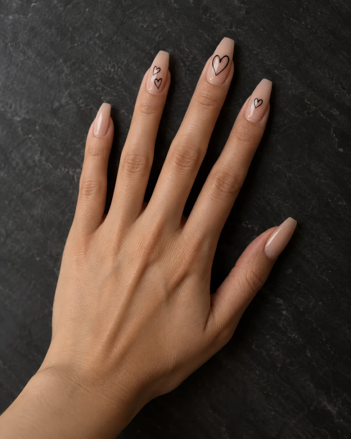Single left hand with coffin nails in warm nude base, thin black outlined hearts at varied sizes on middle, index, and ring fingers, palm down on dark slate