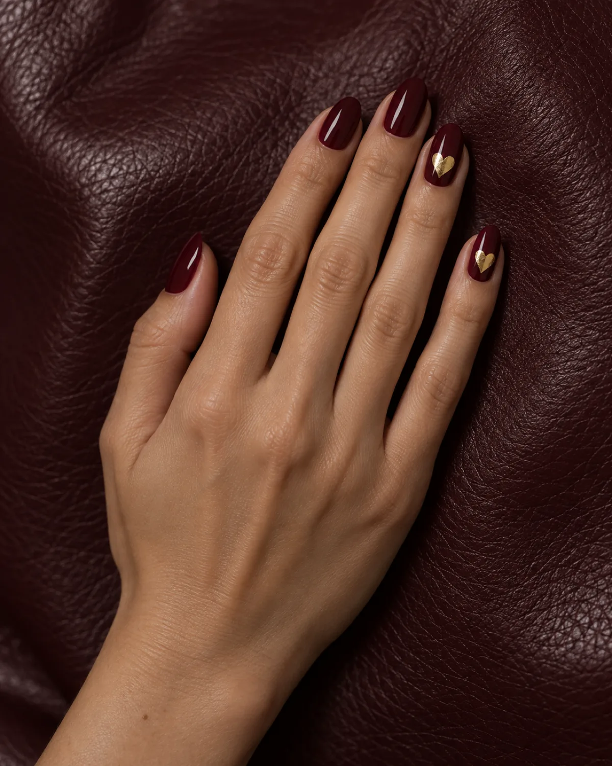 Single right hand with almond nails in rich burgundy crème, ring finger and pinky each with a single irregular gold foil heart with raw imperfect edges, on dark burgundy leather