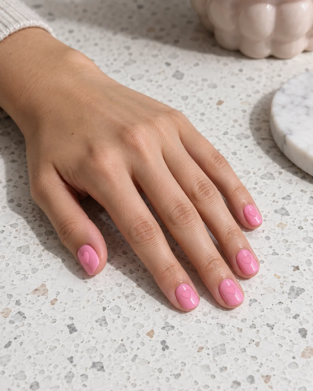 Single left hand with short round nails in glossy bubblegum pink with matte-finish pink hearts in the same hue visible as texture contrast, on white terrazzo