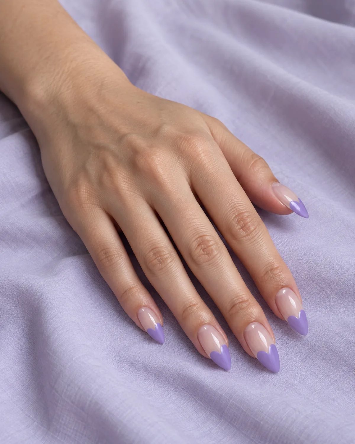 Single left hand with stiletto nails, clear transparent base with only the nail tip painted in lavender-purple as a heart-shaped silhouette, flat on pale lavender fabric