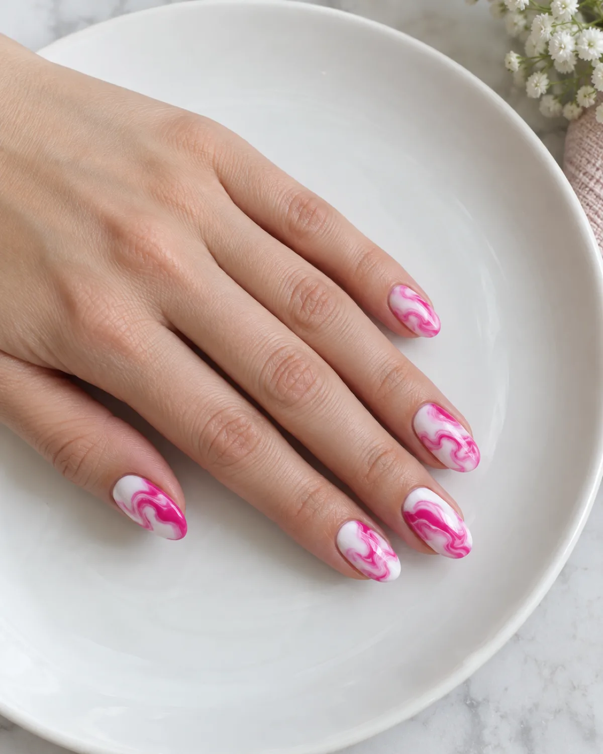 Single left hand with white-and-hot-pink marble swirl nails on medium oval shape resting on a white ceramic plate