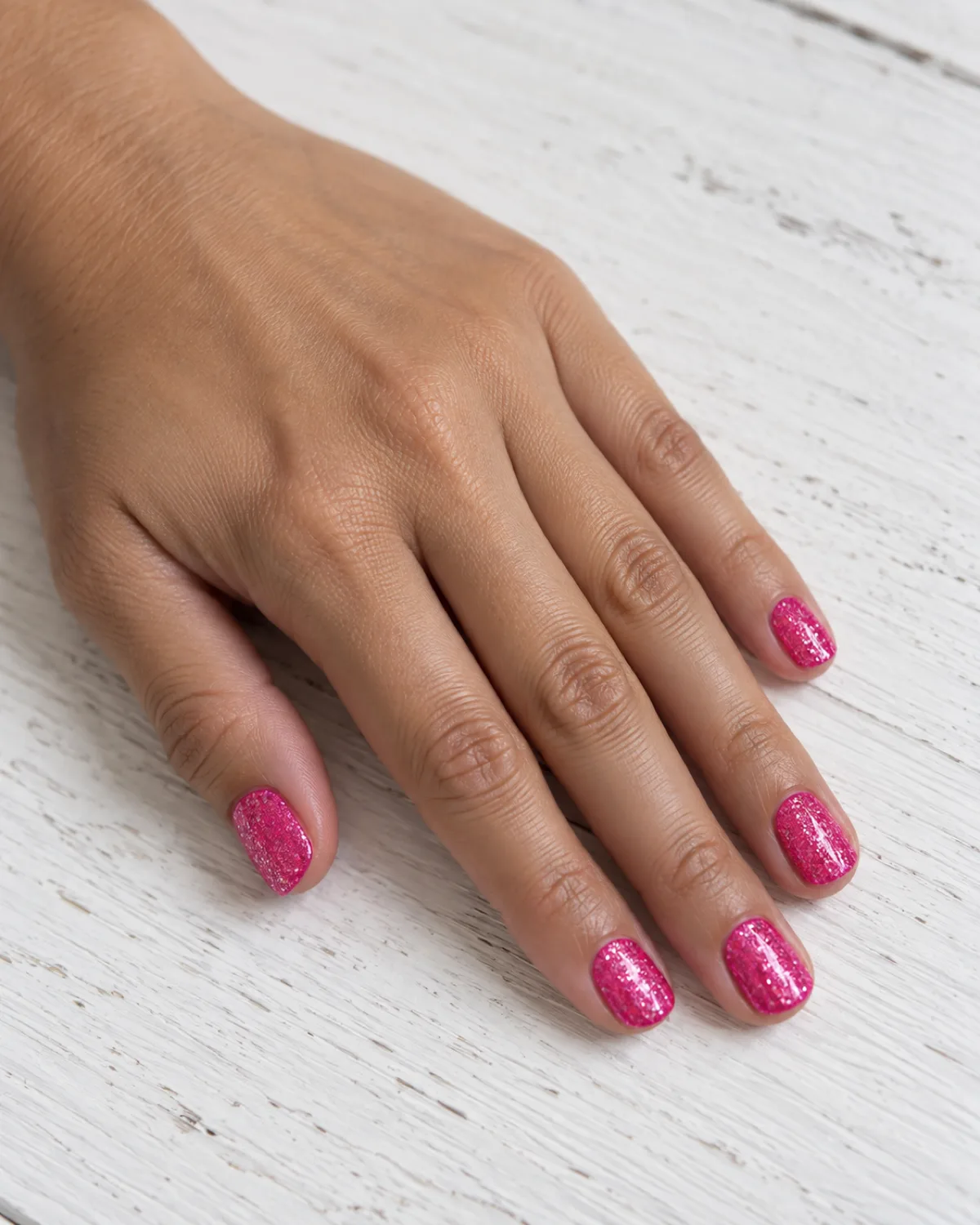 Single left hand with dense hot pink glitter nails on all five fingers on short round shape resting on white wood