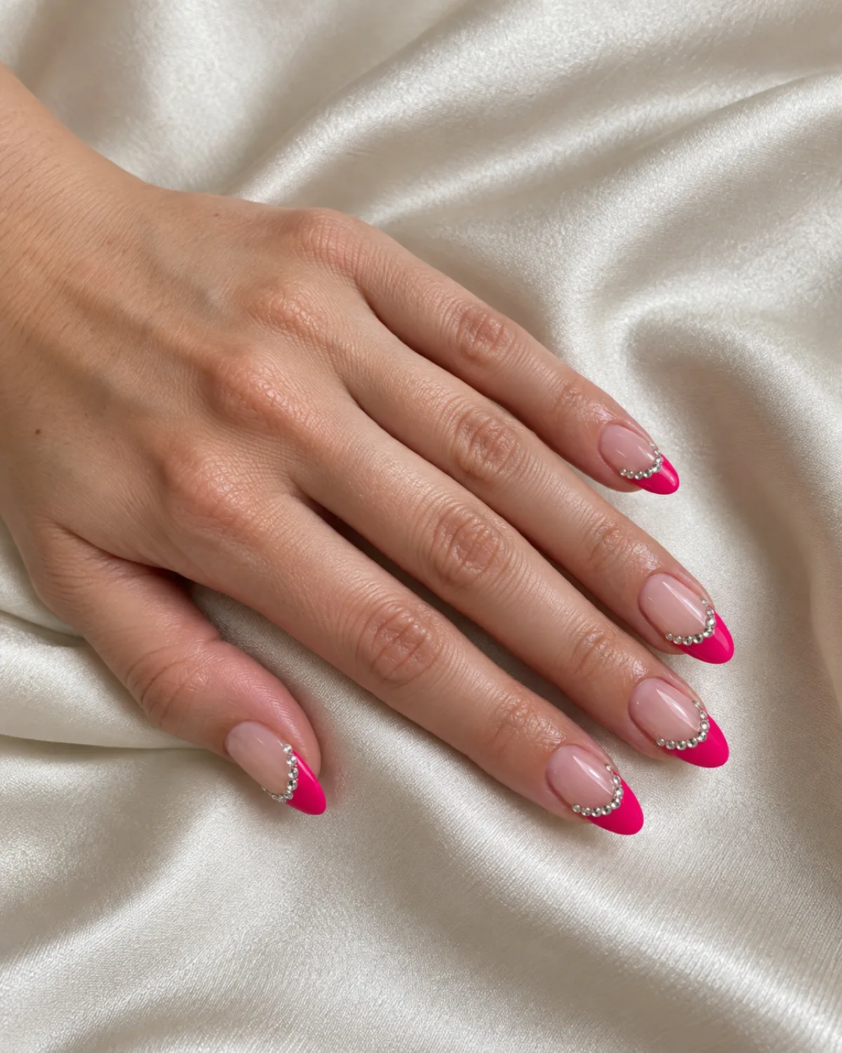 Single left hand with hot pink French tips and tiny crystal gems along the smile line on medium almond nails resting on cream satin