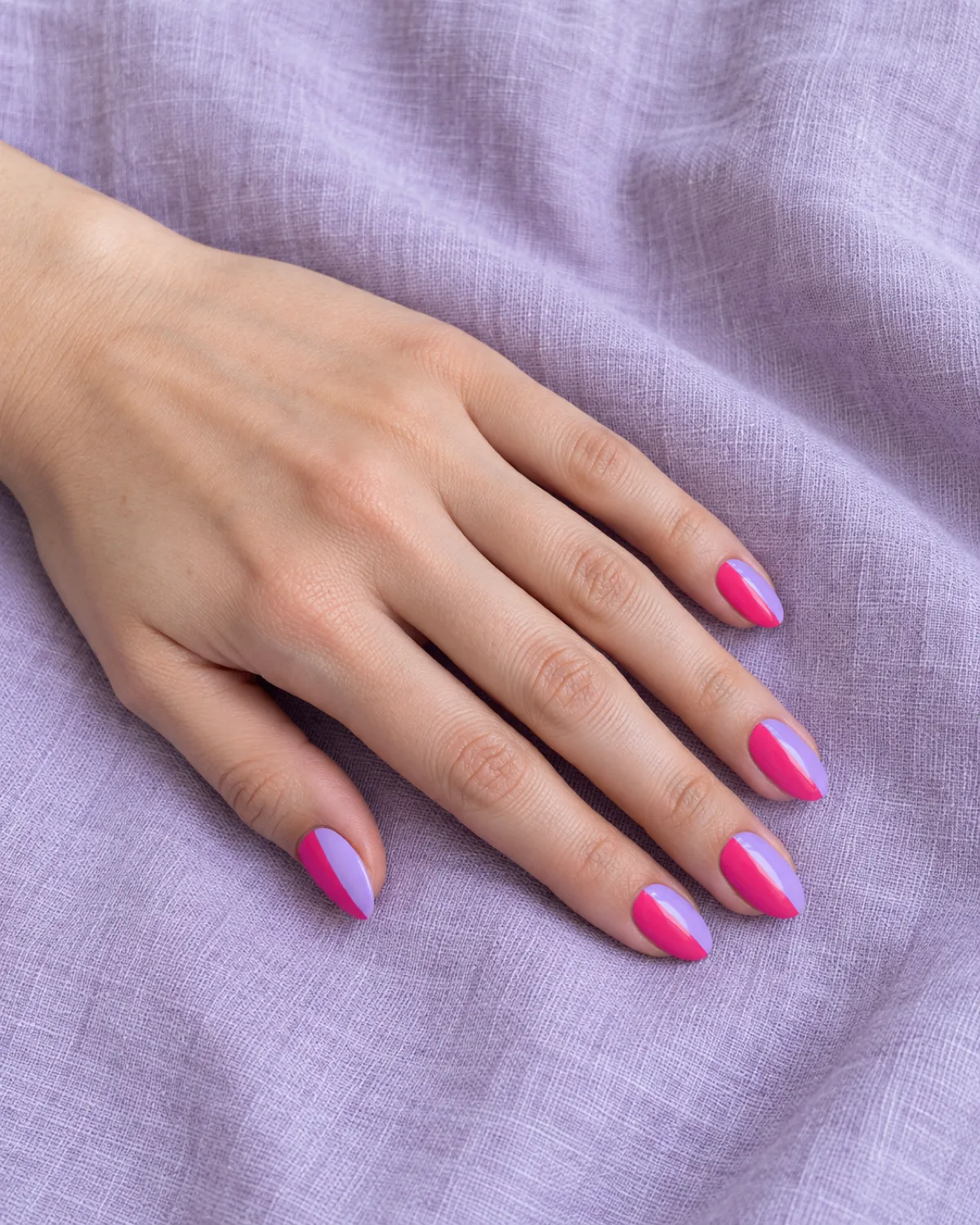 Single left hand with vertically split hot pink and lilac nails on medium almond shape resting on lavender linen