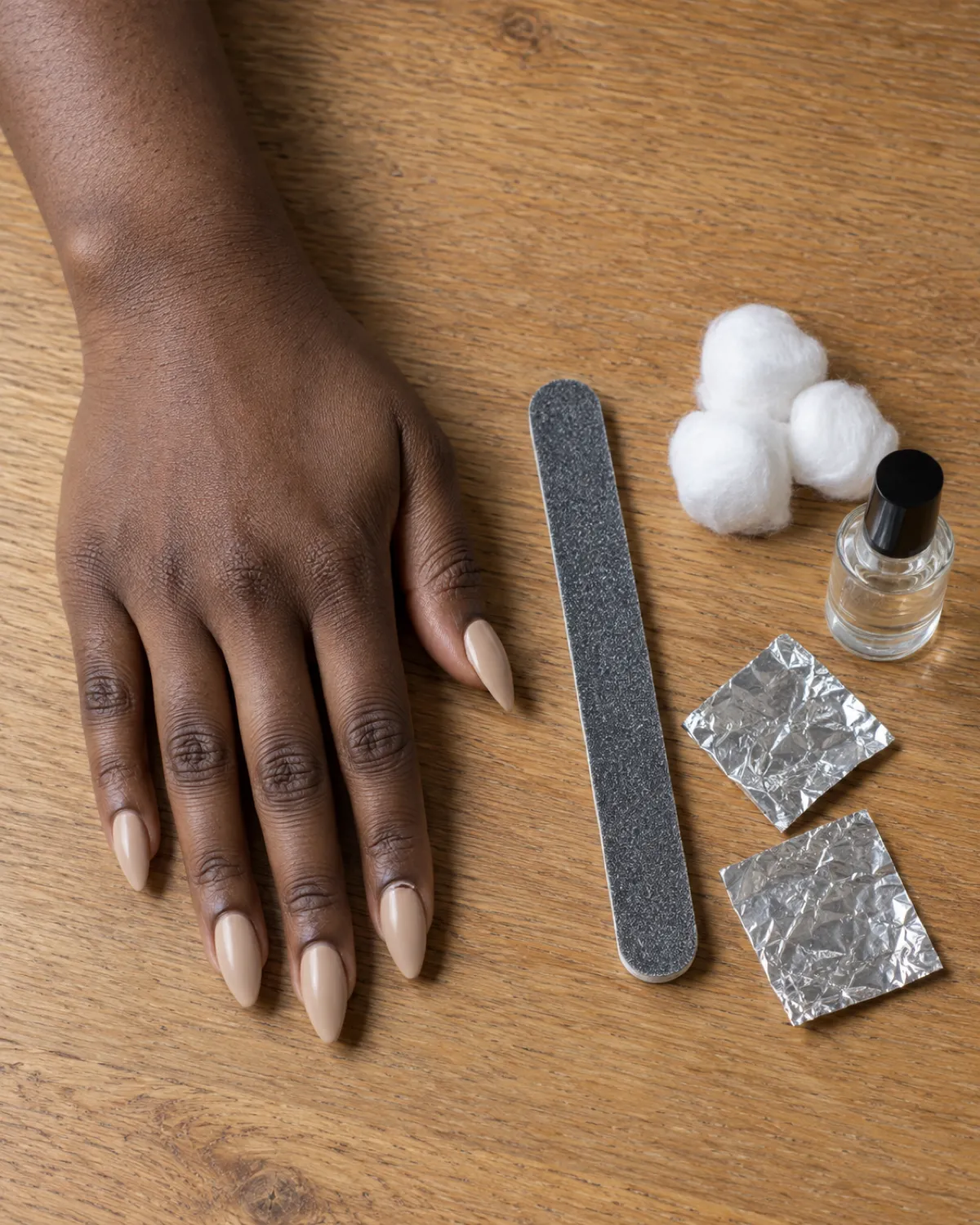 Left hand on oak table with nude acrylic stiletto nails, tools arranged beside it: nail file, cotton balls, aluminium foil squares, and a small glass bottle