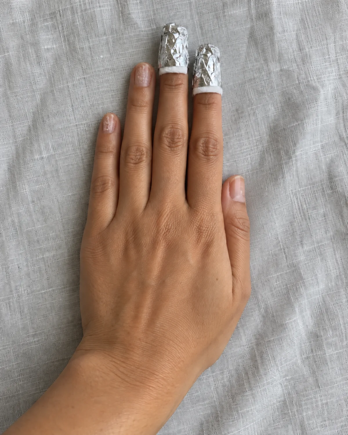 Left hand on grey linen with index and middle fingers wrapped in aluminium foil, cotton edges visible, remaining fingers showing filed acrylic residue