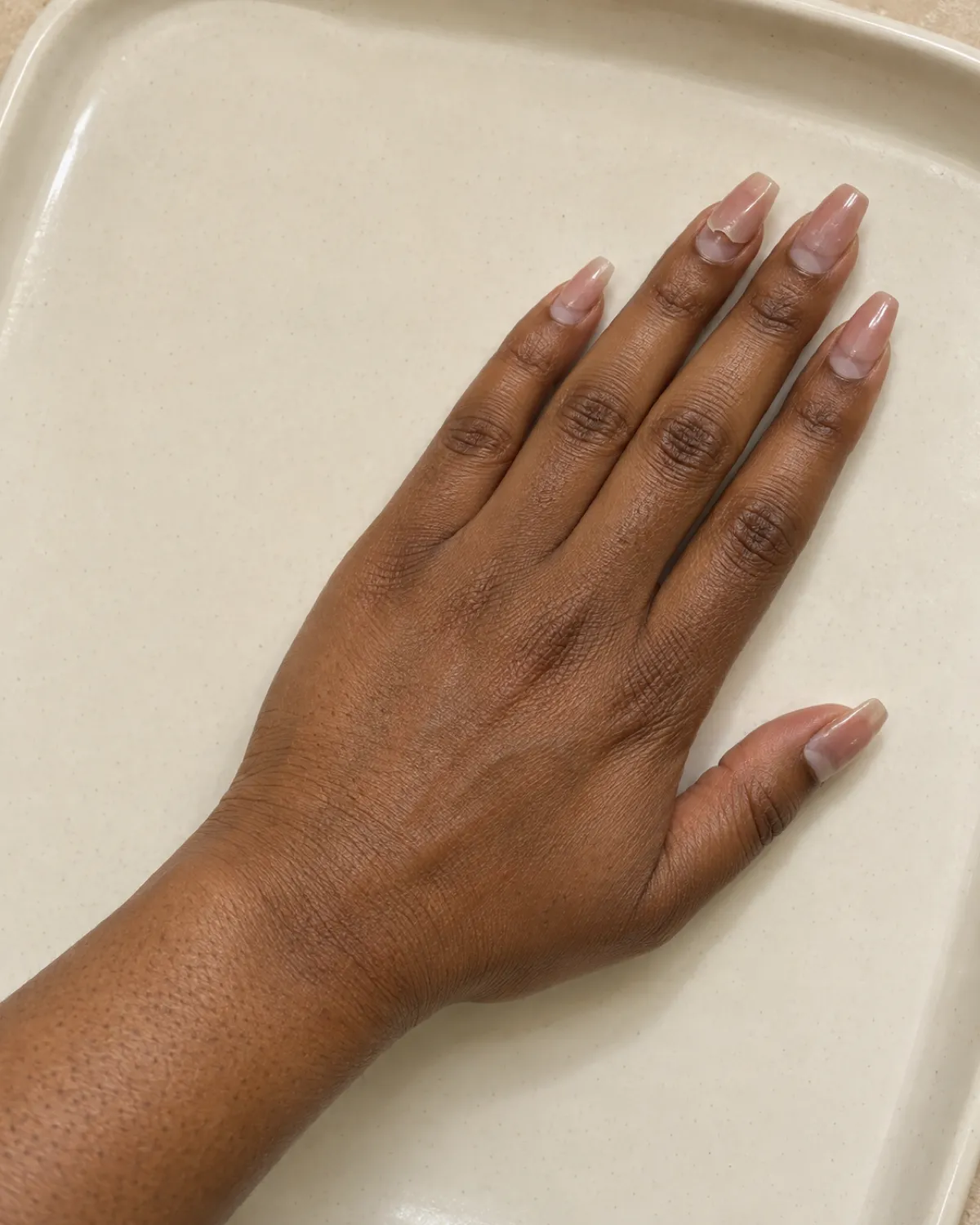 Right hand on a cream ceramic tray showing grown-out acrylic nails with a visible lift at the ring finger cuticle edge