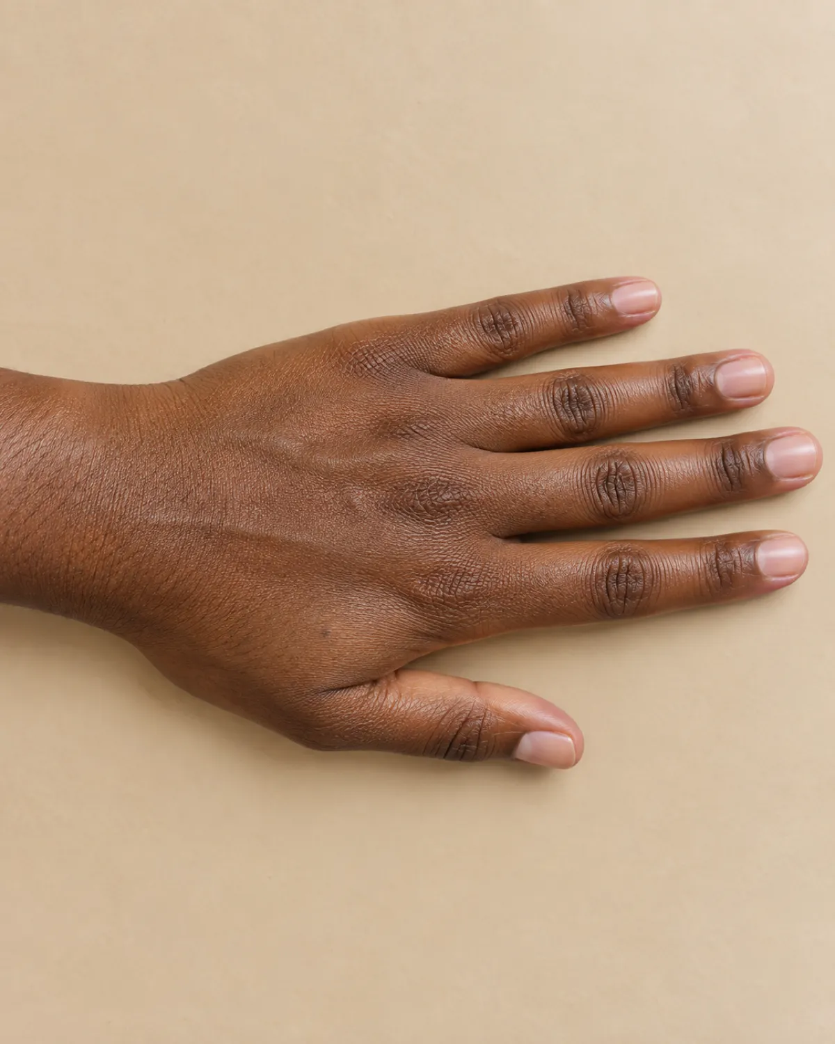Left hand lying flat on warm beige surface with short healthy bare nails showing subtle pink glow and clean cuticle line