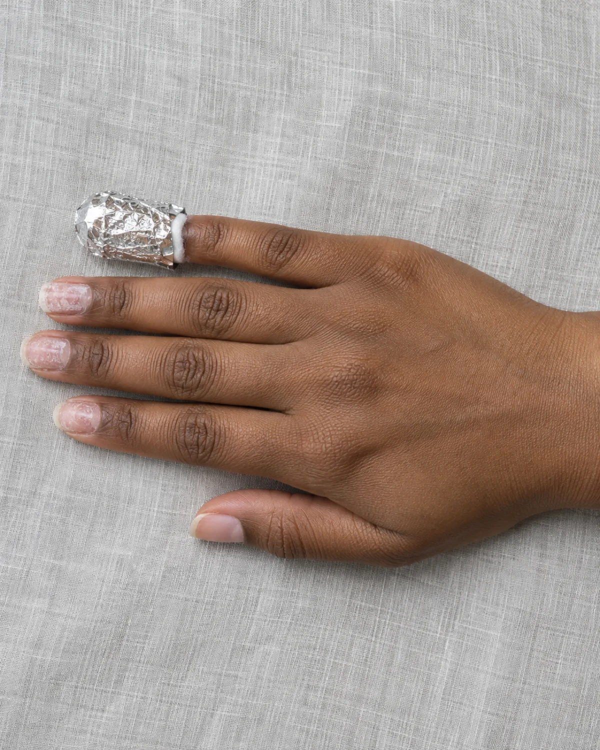 Hand on pale grey linen with the index finger wrapped in aluminium foil showing a cotton edge at the cuticle, other fingers showing partially filed gel residue