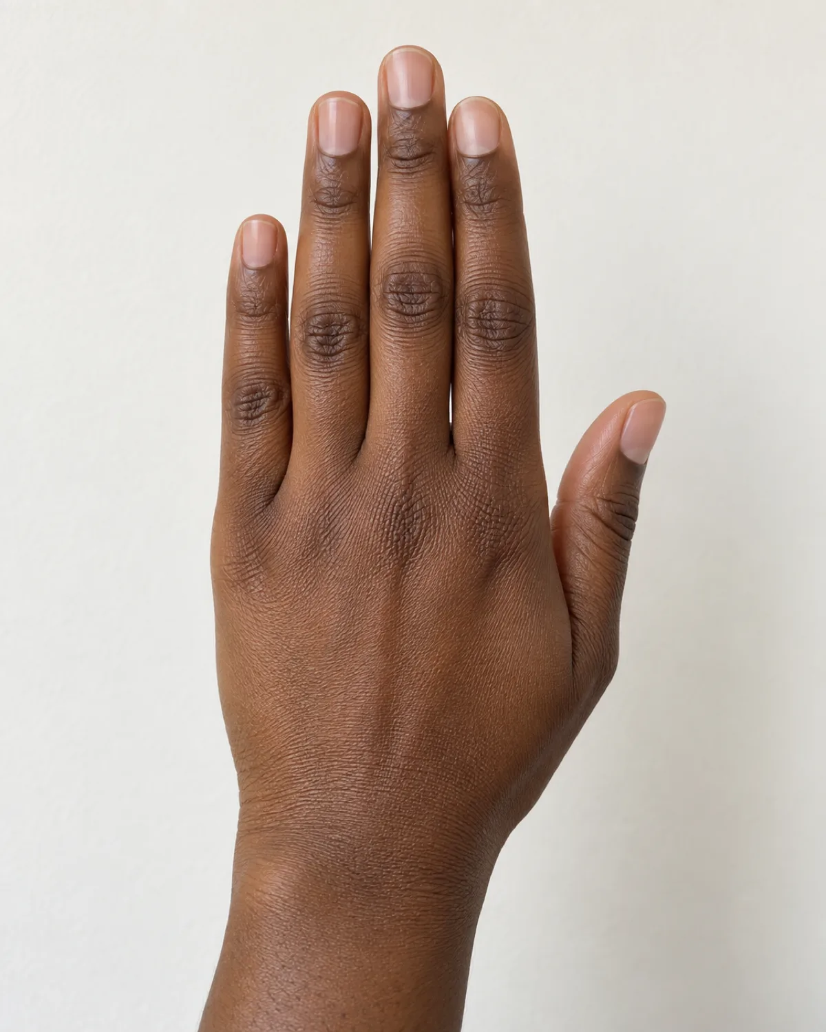 Back of a raised hand with short bare natural nails and a clean cuticle line against a warm white wall, showing freshly cleaned nails after gel removal